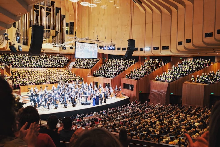 A wide view of the concert hall with a full orchestra on stage, choir in tiered seating, and an audience filling the stalls.