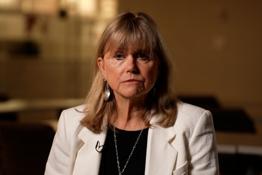 A woman with shoulder-length blonde hair, wearing a light-coloured blazer, sits in an office with a serious expression.