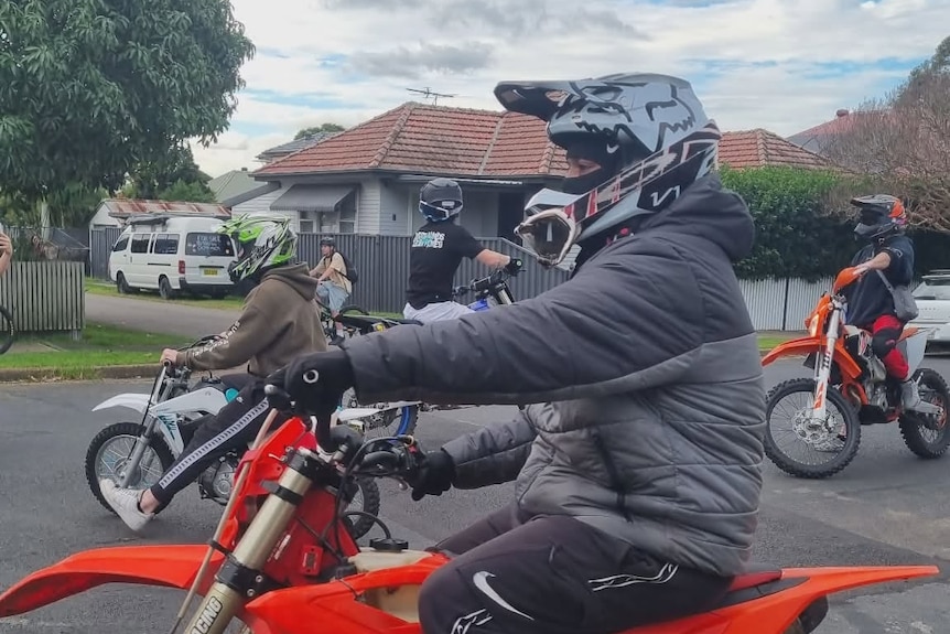 Dirt bike riders wearing helmets riding around a suburban road with houses.