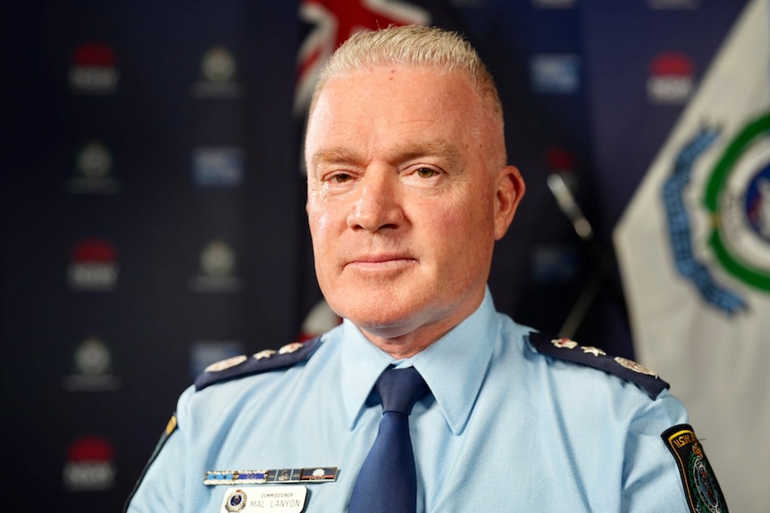 Mal Lanyon close up, in his blue police uniform in front of a flag and NSW Police logo media wall.