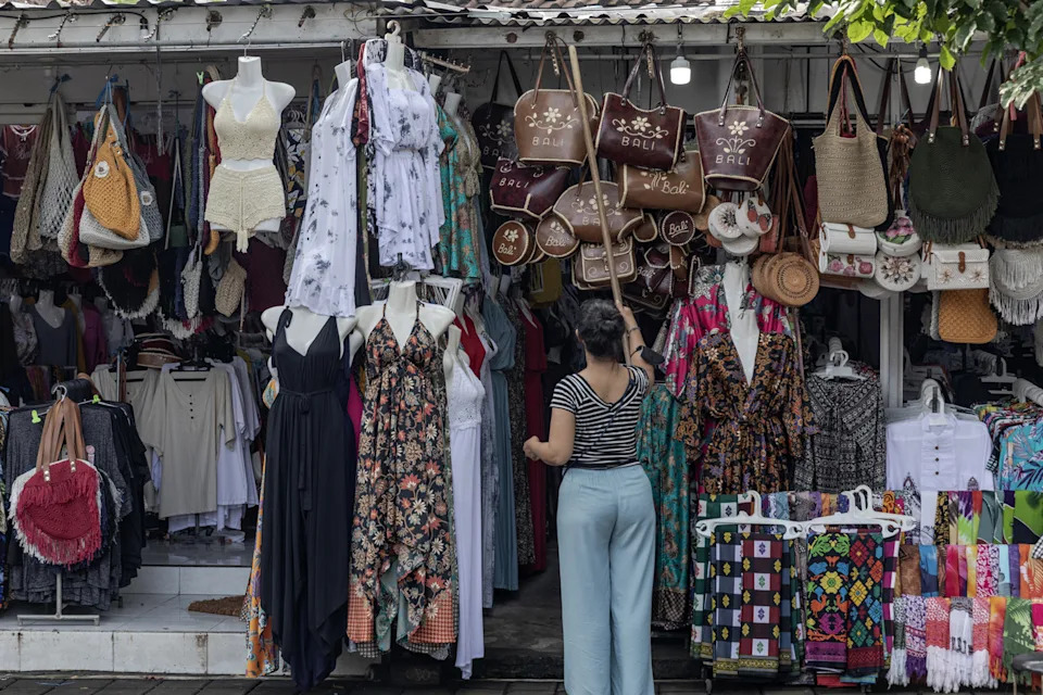 A Bali market selling clothing and accessories, with a woman grabbing handbags with a stick.