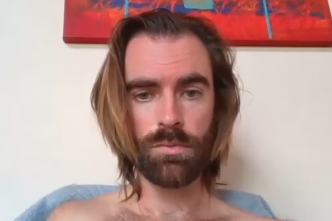 A close-up shot of a shirtless man with long brown hair and a beard, with a weary look while appearing on a video call.