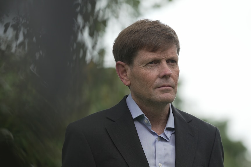 A man wearing a jacket and formal shirt looks to the side of frame.