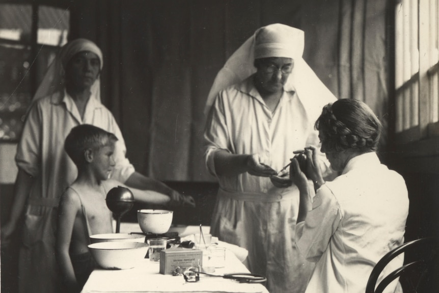A black and white image of a young boy holding out his arm as two women prepare a vaccine dose.