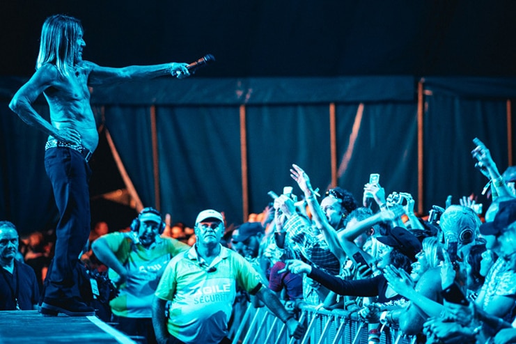 A shirtless older man on a stage holds a microphone over an audience, in blue lighting.