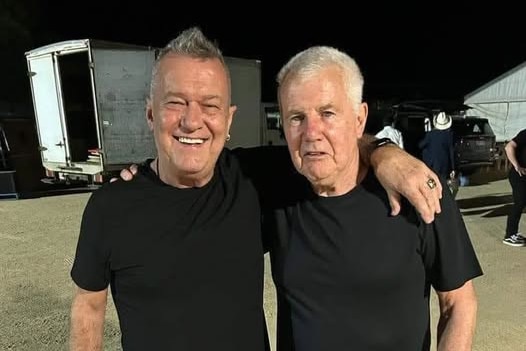 Two grey haired men in black shirts and black jeans stand arm in arm near a truck at night.