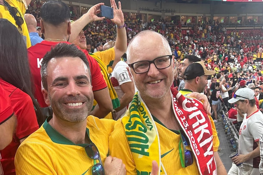 A dark-haired man and a man in glasses smile while sitting among a huge crowd in a football stadium.