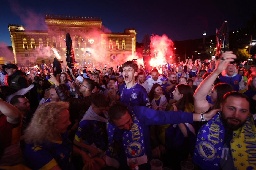 a large crowd at night jump and cheer while smoke flares are set off behind them