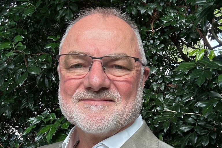 A headshot of a man with grey hair wearing glasses in front of a tree.