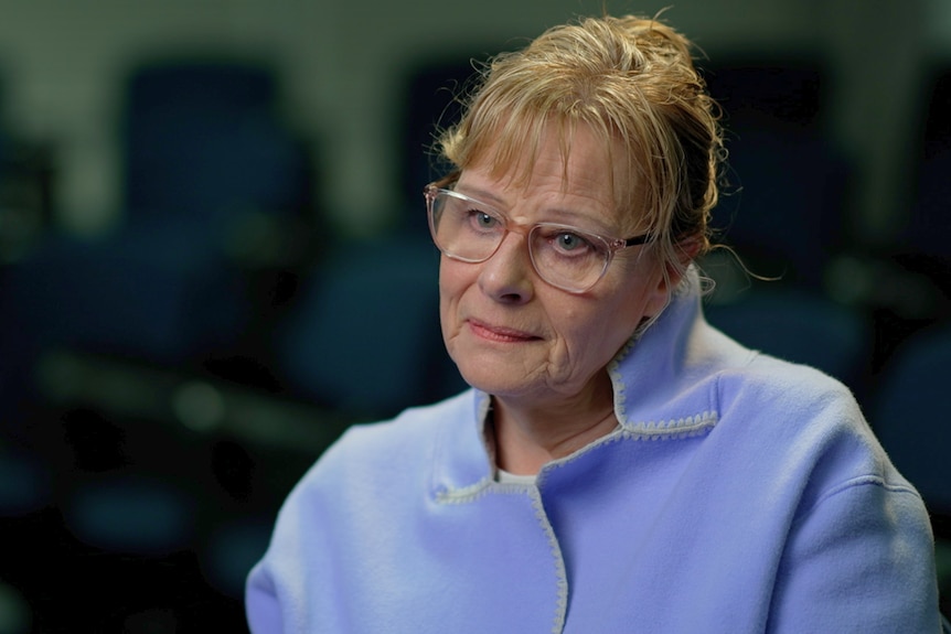 A woman wears a pastel purple blazer and glasses and has short hair.