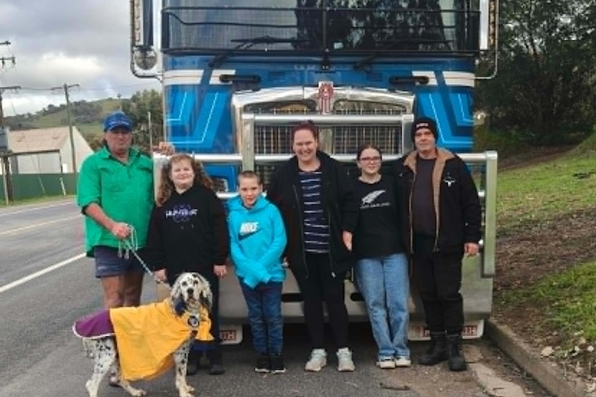 Children, women and a man and a dog stand in front of a parked prime mover.