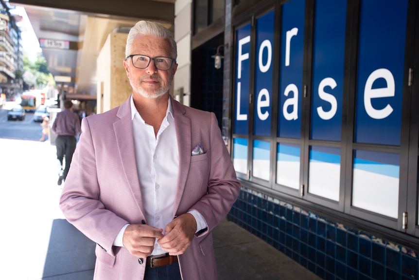 A white-haired man in glasses and a pastel blazer stands next to a building with a "for lease" sign in the window.