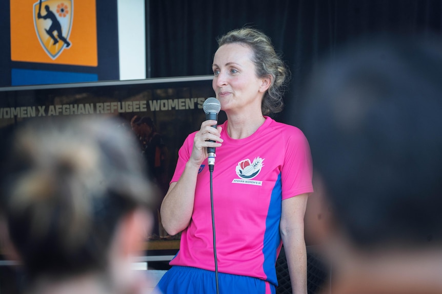 White woman, tied back brown / blonde hair, speaking into microphone in hot pink pink sport polo, soft smile.