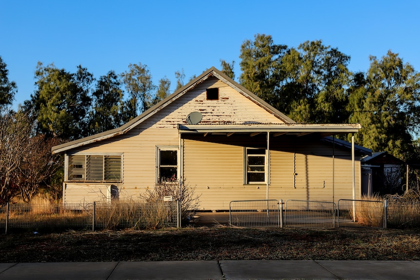 A white weatherboard house bathed in golden light, paint is worn in places, glass is missing from windows.