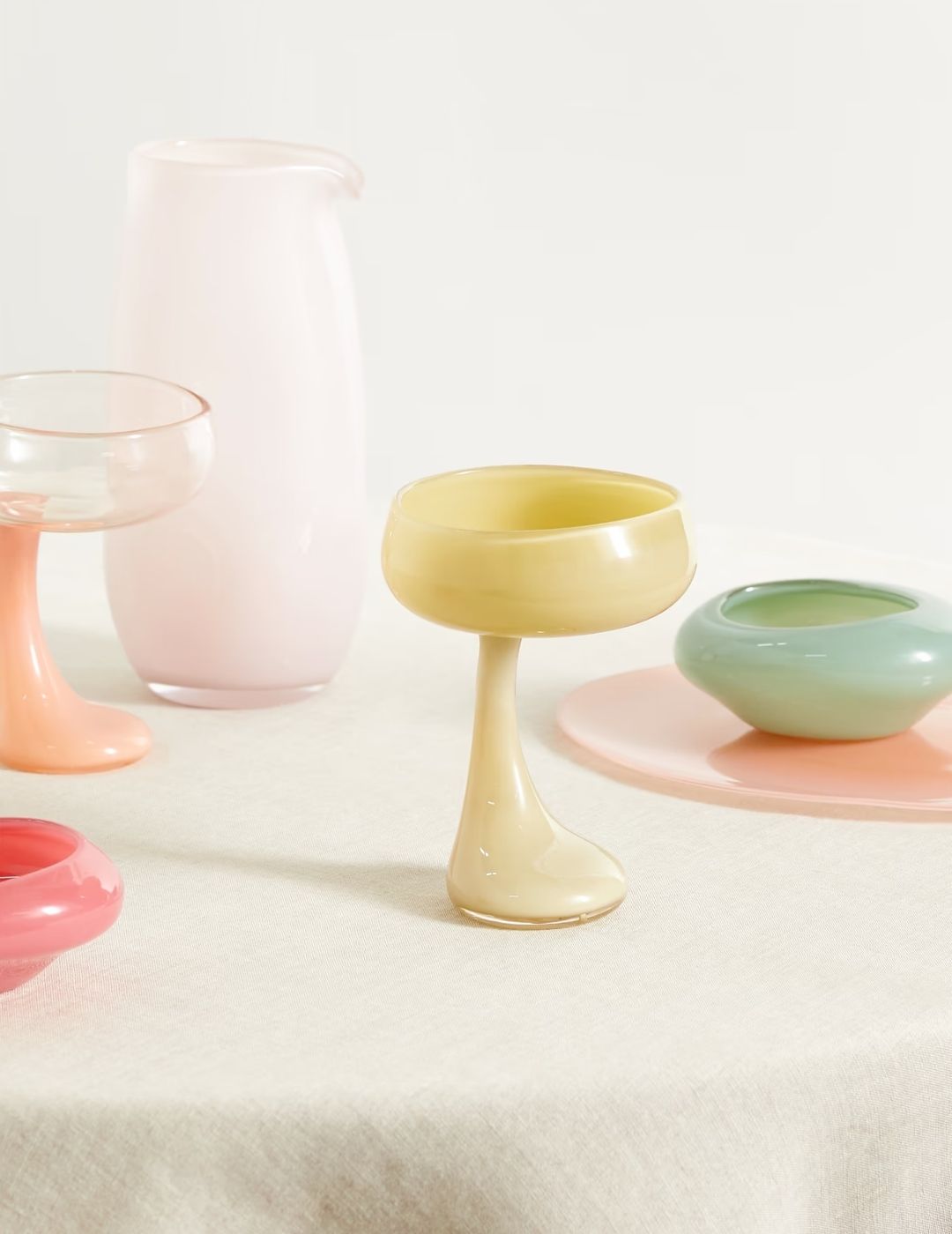 A series of mouth-blown glass pieces, including cups, plates, and a vase, sit on a beige tablecloth.