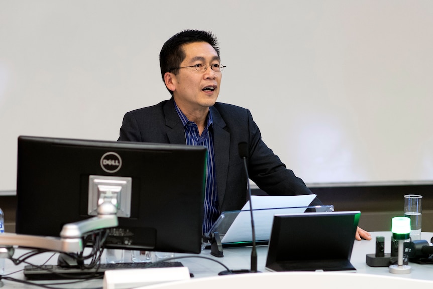Professor Tele tan speaks into a microphone in a lecture hall, wearing a suit jacket and dark blue collared shirt.
