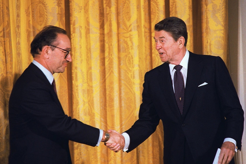 Alan Greenspan (left) shakes Ronald Reagan's hand while they wear suits.