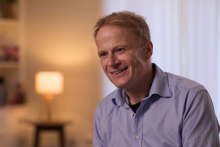 A man with wispy short blonde hair wears a blue collared shirt, seated, he is smiling. A lamp on behind him