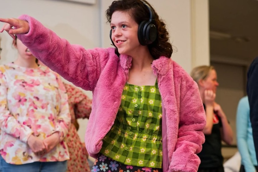 A teenage girl dancing. She is wearing headphones, a pink jacket and green top.