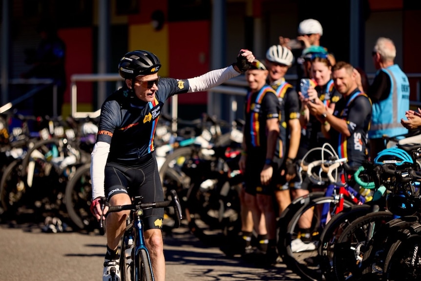 Man on a bicycle with helmet and black riding gear waves to an audience of people. Bikes lined up in the background