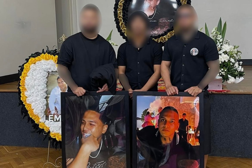 Three people with faces and tattooed arms blurred stand behind large image of man with shaved head, flowers behind them