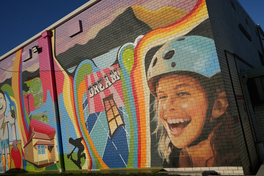 Colourful mural on a brick wall next to skate park, showing girl smiling wide wearing a blue helmet.