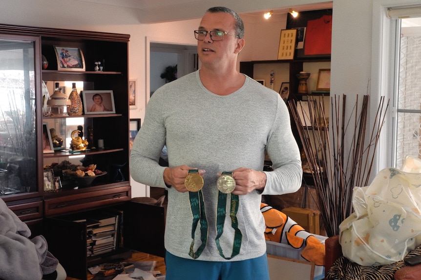 A man wearing a long sleeved grey shirt and glasses holds a silver and bronze medal standing in his living room.
