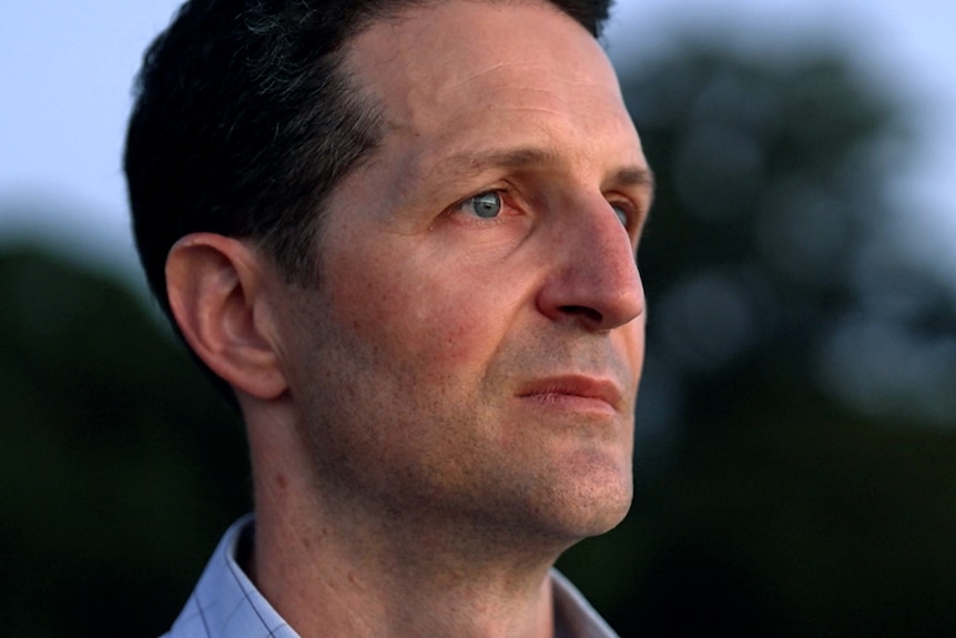 A close-up of Kurt Hayward's face. He is standing outdoors, looking straight ahead with a serious, slightly sad expression.