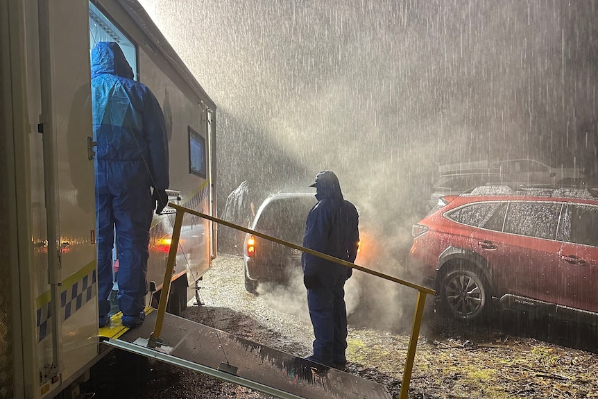 Police investigators in blue forensic crime scene suits stand in heavy rain at night