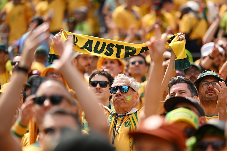 in a sea of australian fans on man holds up a green and gold scarf that says 'Australia'
