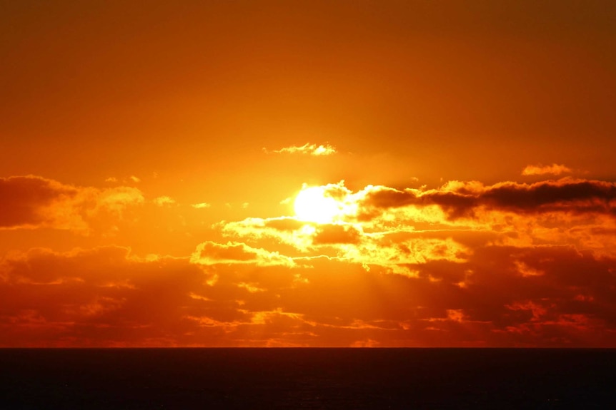 A bright orange sun sets behind clouds over a beach.