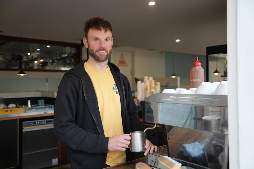 A man with brown hair in a yellow shirt and black jacket holds a silver jug under a milk frother on a coffee machine.