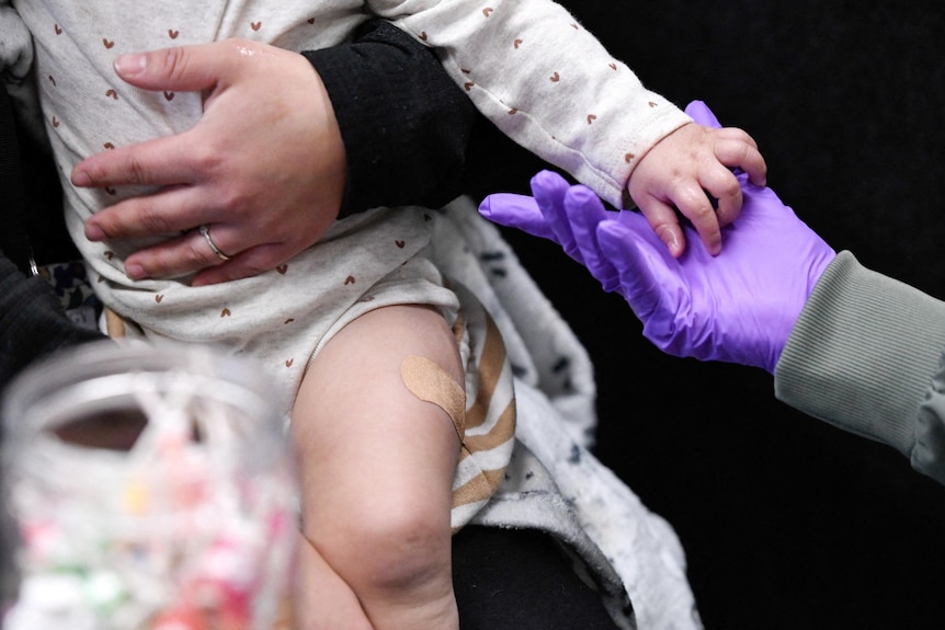 A close up of an infant on their parent’s lap with a band-aid on their left thigh after a vaccine.