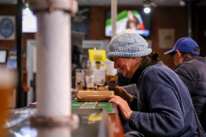 A woman wearing a light blue beanie and blue jumper leans on the wooden bar at a pub. There are beer taps in the foreground.
