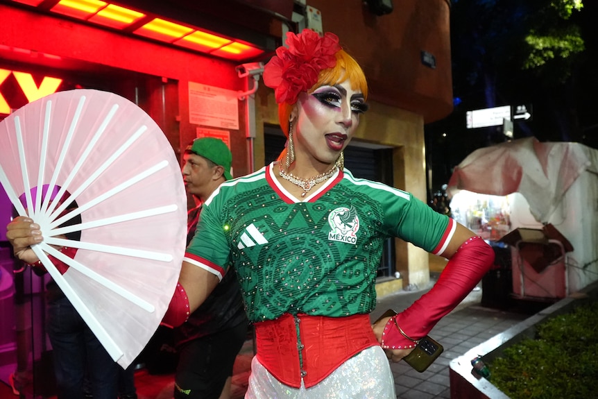 A drag queen wearing a Mexico football jersey, holds a fan and poses.