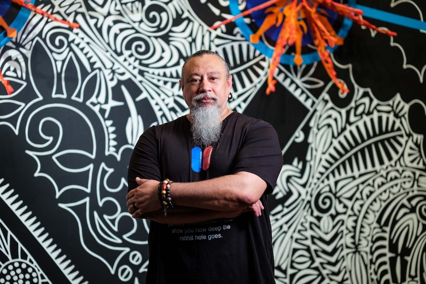 A Torres Strait Islander man with a grey beard standing with his arms crossed in front of a large artwork.
