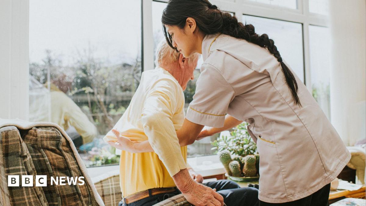 A young female carer helps an elderly male to get dressed by pulling on his jumper, as he sits in an armchair.