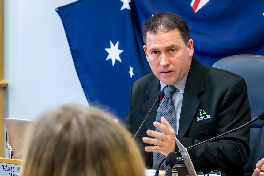 A man behind a desk with a flag in the background looking at someone off camera