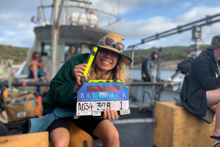 A woman with a clapper board on a boat on a film set.