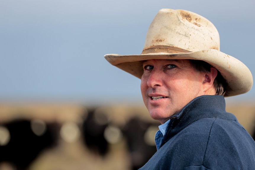 Man wearing weathered wide brim hat looks over shoulder with cattle out of focus in background on flat plains
