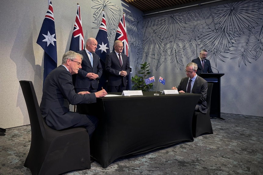 Anthony Albanese and Christopher Luxon standing by a dark table as two men sign documents.