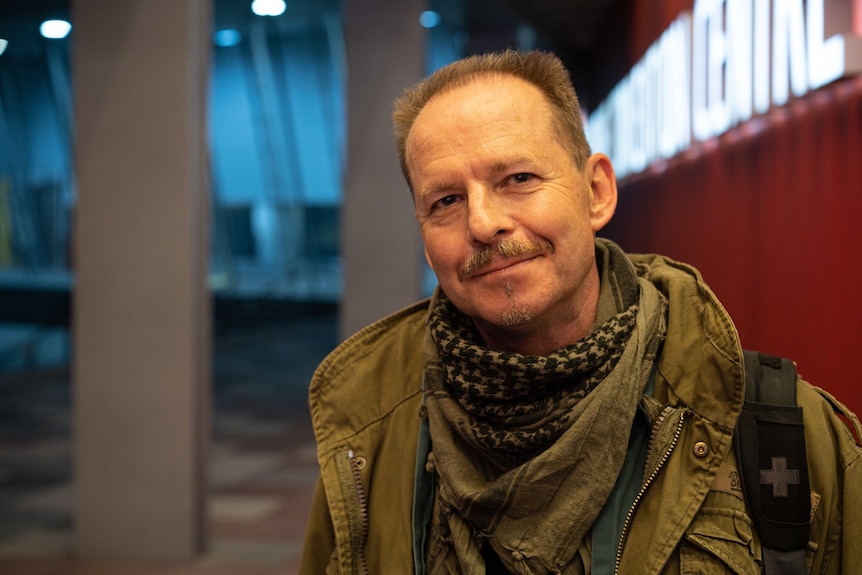 Tight shot of a man with short brown hair in a scarf and green jacket looking into the camera.