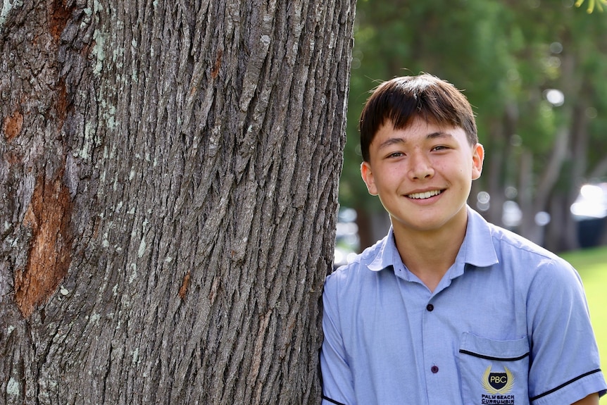 A high school boy leans against a tree