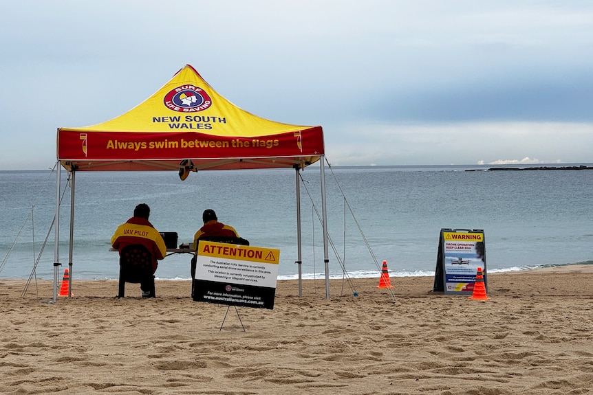 A surf life saving tent with two people in uniform under it with signs saying drone surveillance is happening.