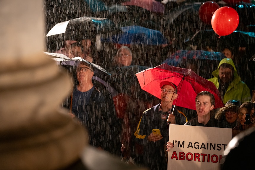 A crowd in pouring rain with umbrellas. One holds a sign saying "I'm against abortions".