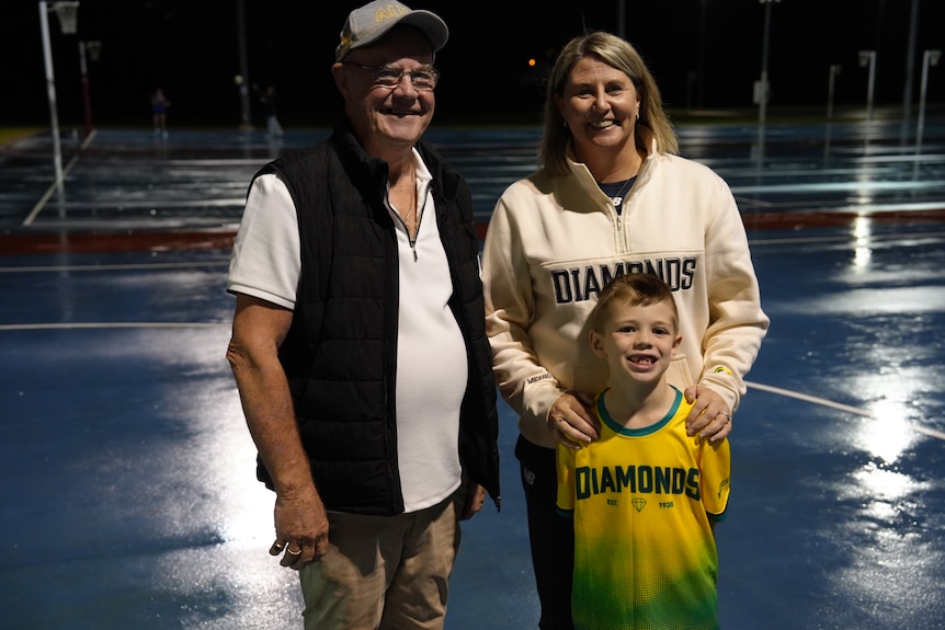 Stacey Marinkovich poses for a photo at outdoor netball courts at night time with son and dad.