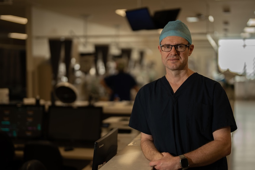 Man wearing hospital scrubs, standing in a hospital ward, looking at camera.