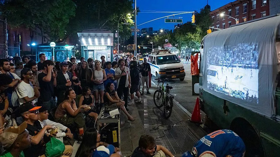 dozens of people gather on a pavement to watch a Knicks game projected onto a sheet on a vehicle