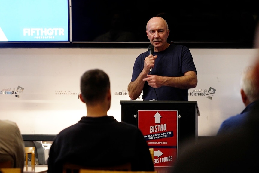 A man in a t-shirt hold a mic and stands behind a lectern, giving a presentation to a group.