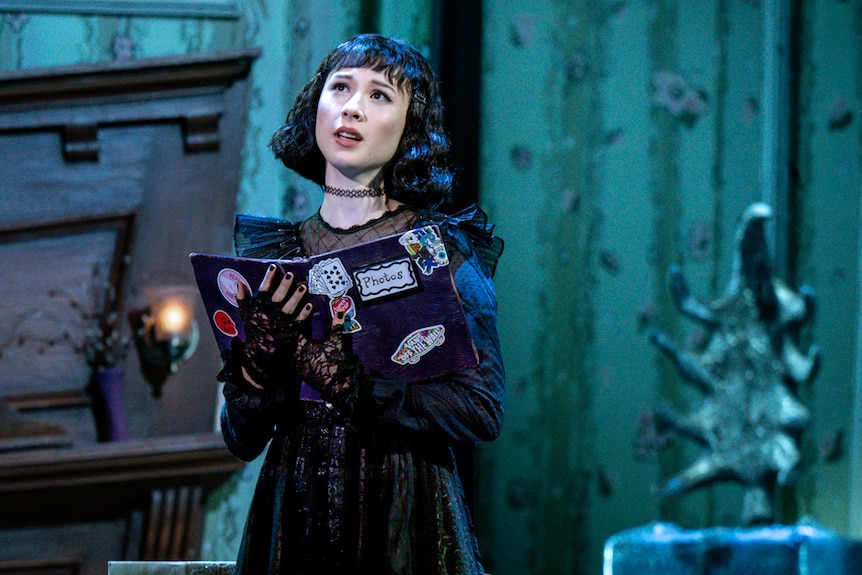 On stage, Karis Oka, a young woman, dressed as a goth teenager, holds a book in her hands as she sings.